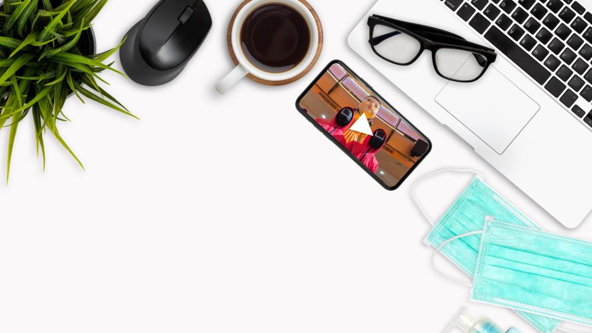 white desk seen from above, with a laptop, phone, glasses, coffee cup, mouse, plant, and face masks