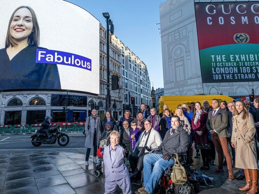 People standing in Piccadilly Circus posing in front of Purple Tuesday digital ad on a big screen